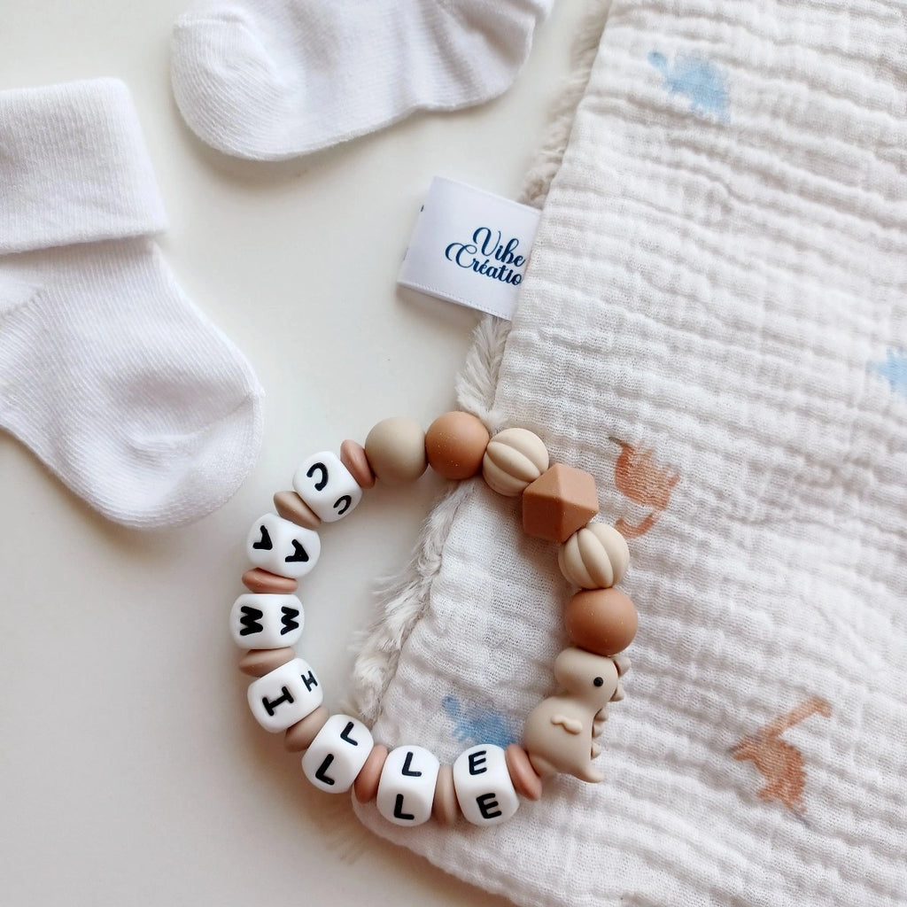 Anneau de dentition pour soulager les gencives des bébés. Cet anneau de dentition en forme de bracelet, est En silicone alimentaire sans BPA, entièrement personnalisables, perles beiges et marron, perles du prénom et dinosaure couleur taupe. Idéal cadeau naissance