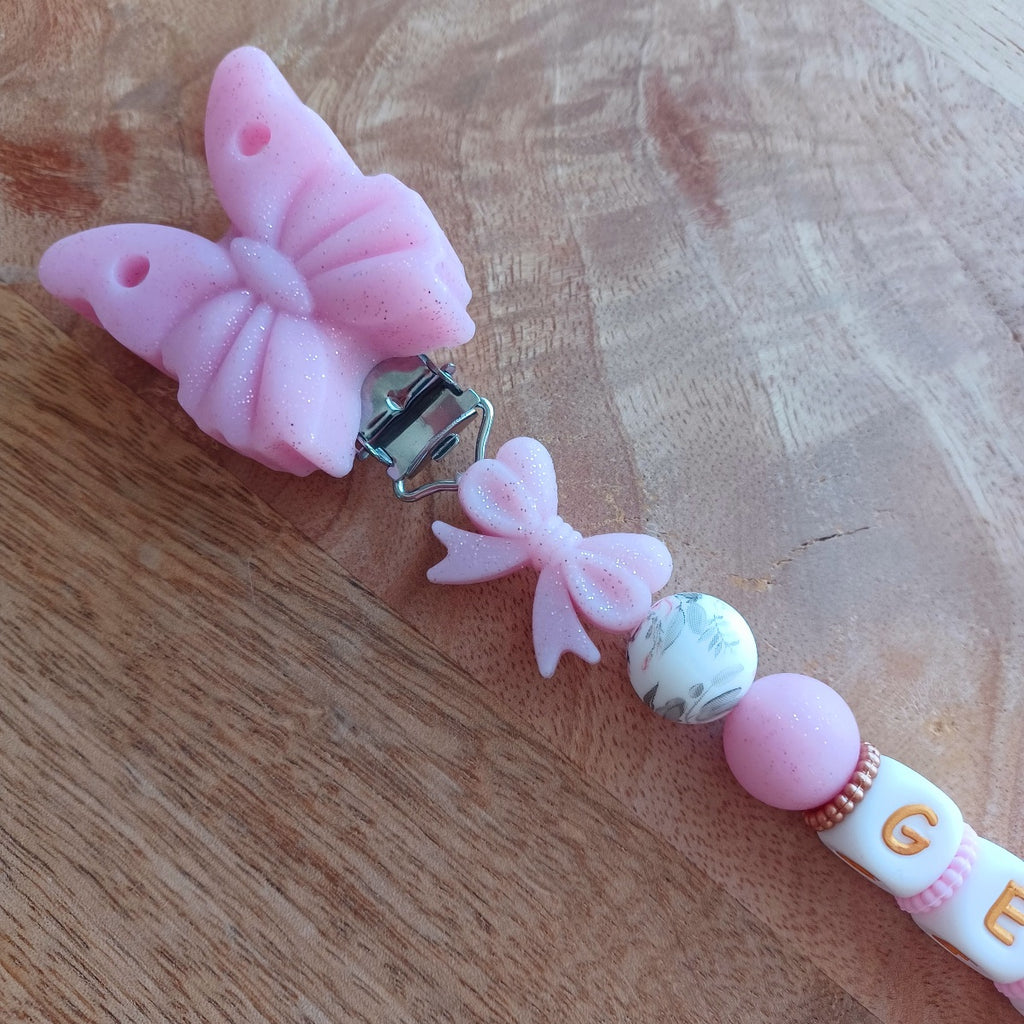 Attache tétine en silicone alimentaire sans BPA. Personnalisable avec le prénom du bébé, perles rondes, perles imprimées, perles en forme de fleurs, de noeuds, perles pailletées. Clip en silicone forme papillon. Cadeau de naissance