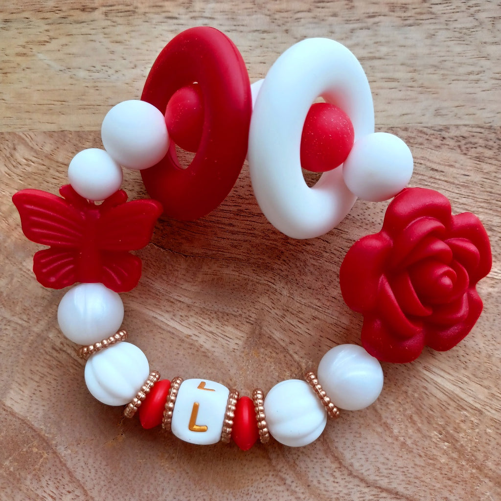 Attache tétine et anneau de dentition en silicone alimentaire sans BPA.  Clip rond en silicone et acier inoxydable, perles prénom personnalisable, papillon rouge en silicone. Anneau de dentition en taille parfaite pour les petites mains du bébé. Cadeau de naissance