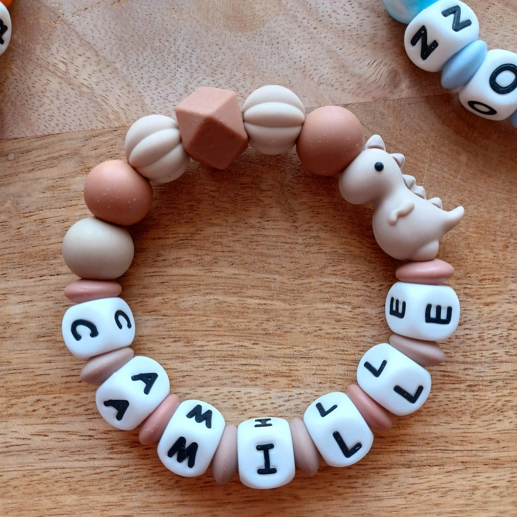 Anneau de dentition pour soulager les gencives des bébés. Cet anneau de dentition en forme de bracelet, est En silicone alimentaire sans BPA, entièrement personnalisables, perles beiges et marron, perles du prénom et dinosaure couleur taupe. Idéal cadeau naissance