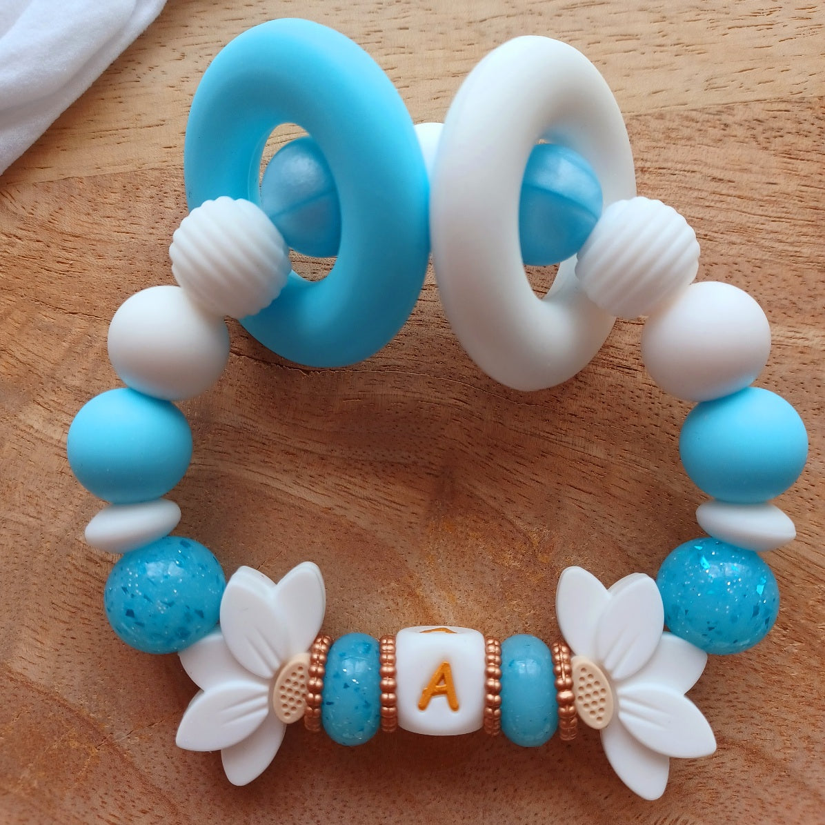 Attache tétine et anneau de dentition personnalisables avec le prénom de l'enfant. Réalisé en silicone alimentaire sans BPA, non toxique. Perles pailletées, perles en forme de fleur de lotus, clip en forme de coeur, pince en acier inoxydable. Super idée cadeau, cadeau de naissance. Couleur bleu et blanc, lettre en couleur blanc et doré.