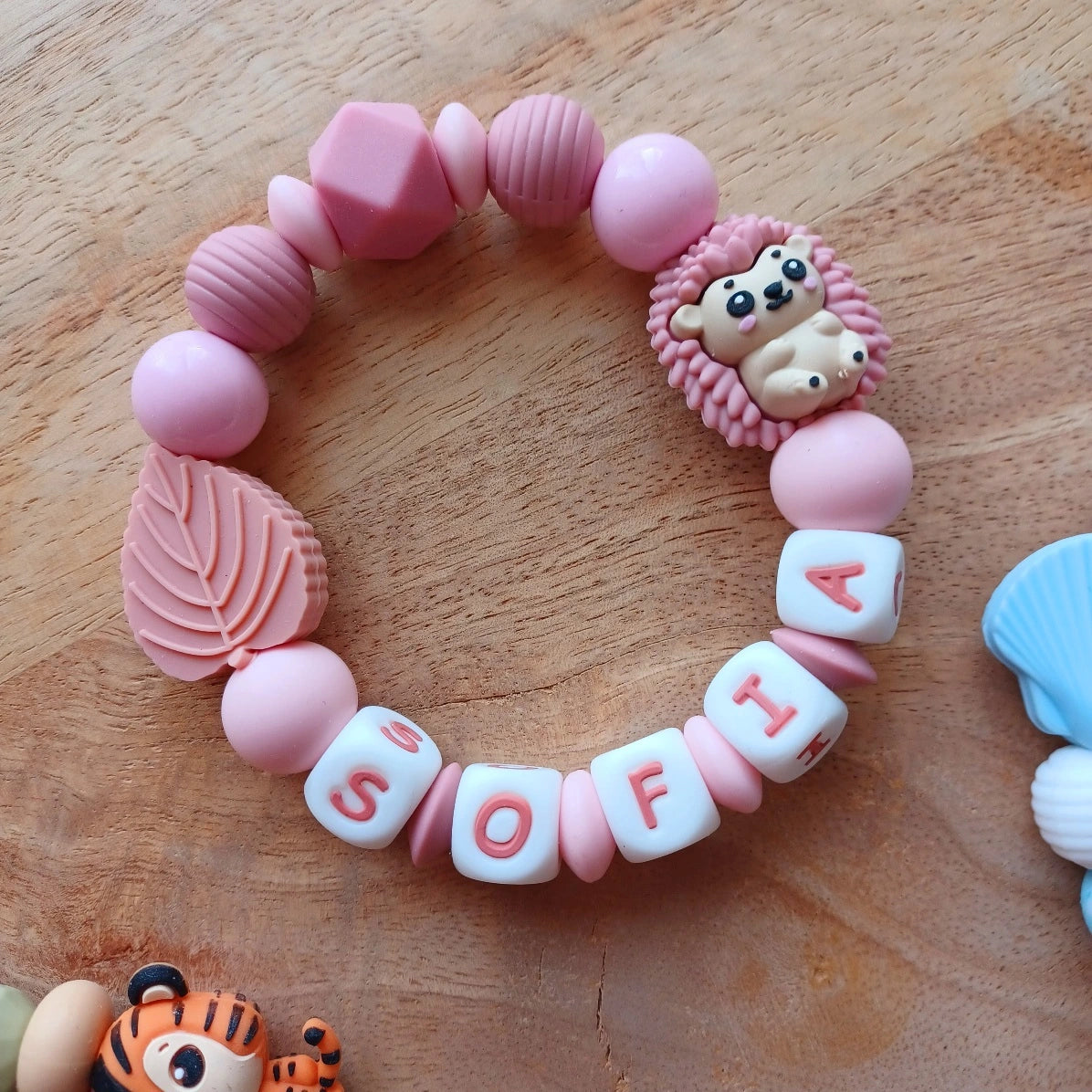 Anneau de dentition pour soulager les gencives des bébés. Cet anneau de dentition en forme de bracelet, est En silicone alimentaire sans BPA, entièrement personnalisables, perles rose, rose poudré, vieux rose, perles du prénom et perles fantaisie en forme d'hérison, et feuille. Idéal cadeau naissance