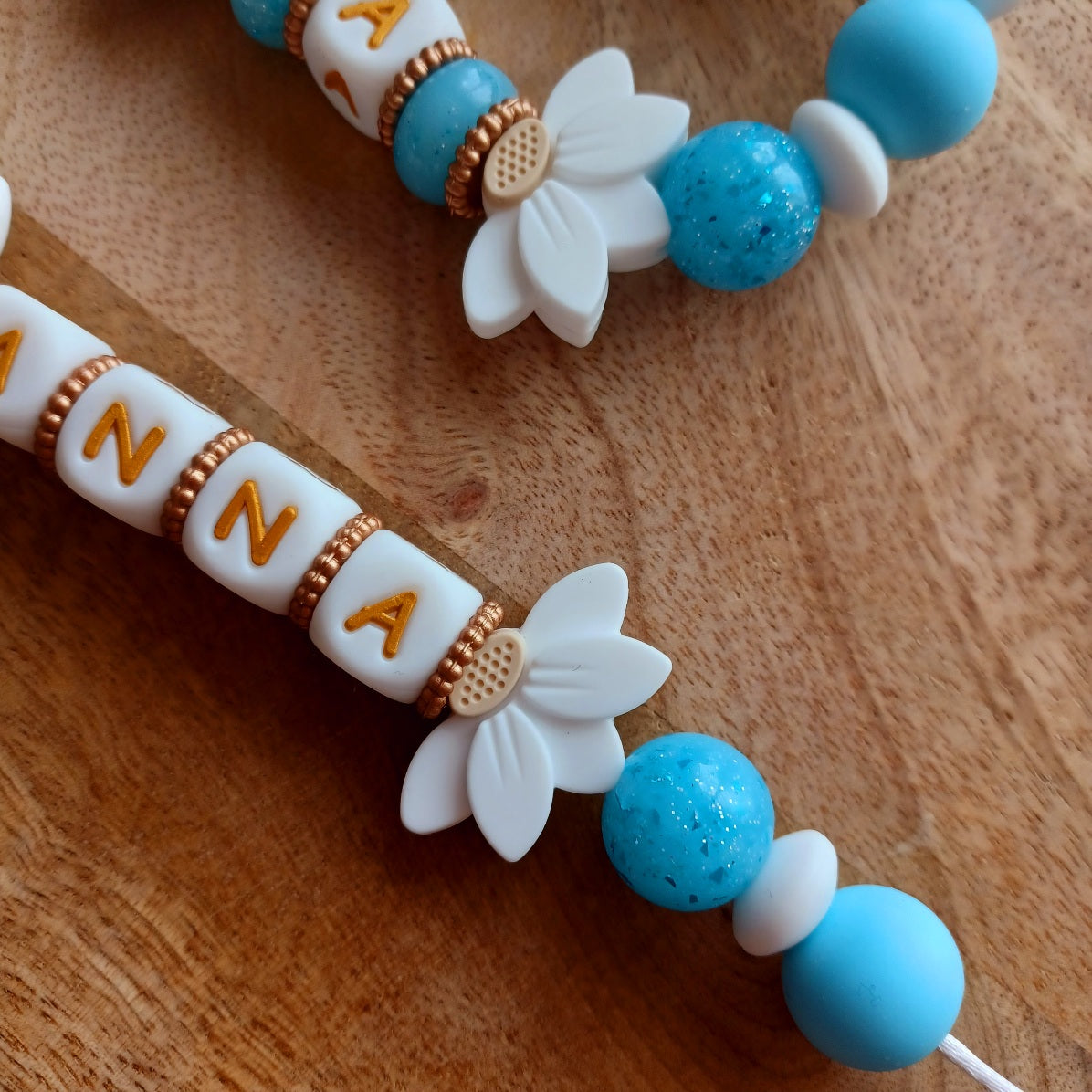 Attache tétine et anneau de dentition personnalisables avec le prénom de l'enfant. Réalisé en silicone alimentaire sans BPA, non toxique. Perles pailletées, perles en forme de fleur de lotus, clip en forme de coeur, pince en acier inoxydable. Super idée cadeau, cadeau de naissance. Couleur bleu et blanc, lettre en couleur blanc et doré.