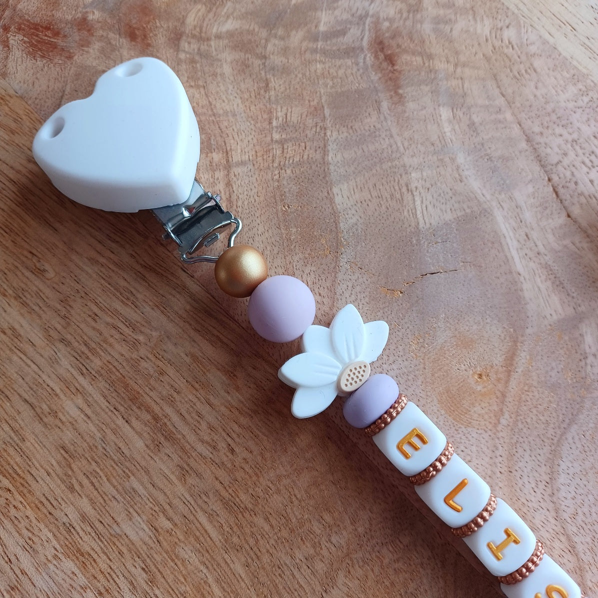 Attache tetine personnalisable, avec prénom du bébé ou enfant. Clip forme de coeur en silicone et acier inoxydable, perles du prénom en blanc et doré, perles rondes en lilas, perles de fleur de lotus. Cadeau de naissance