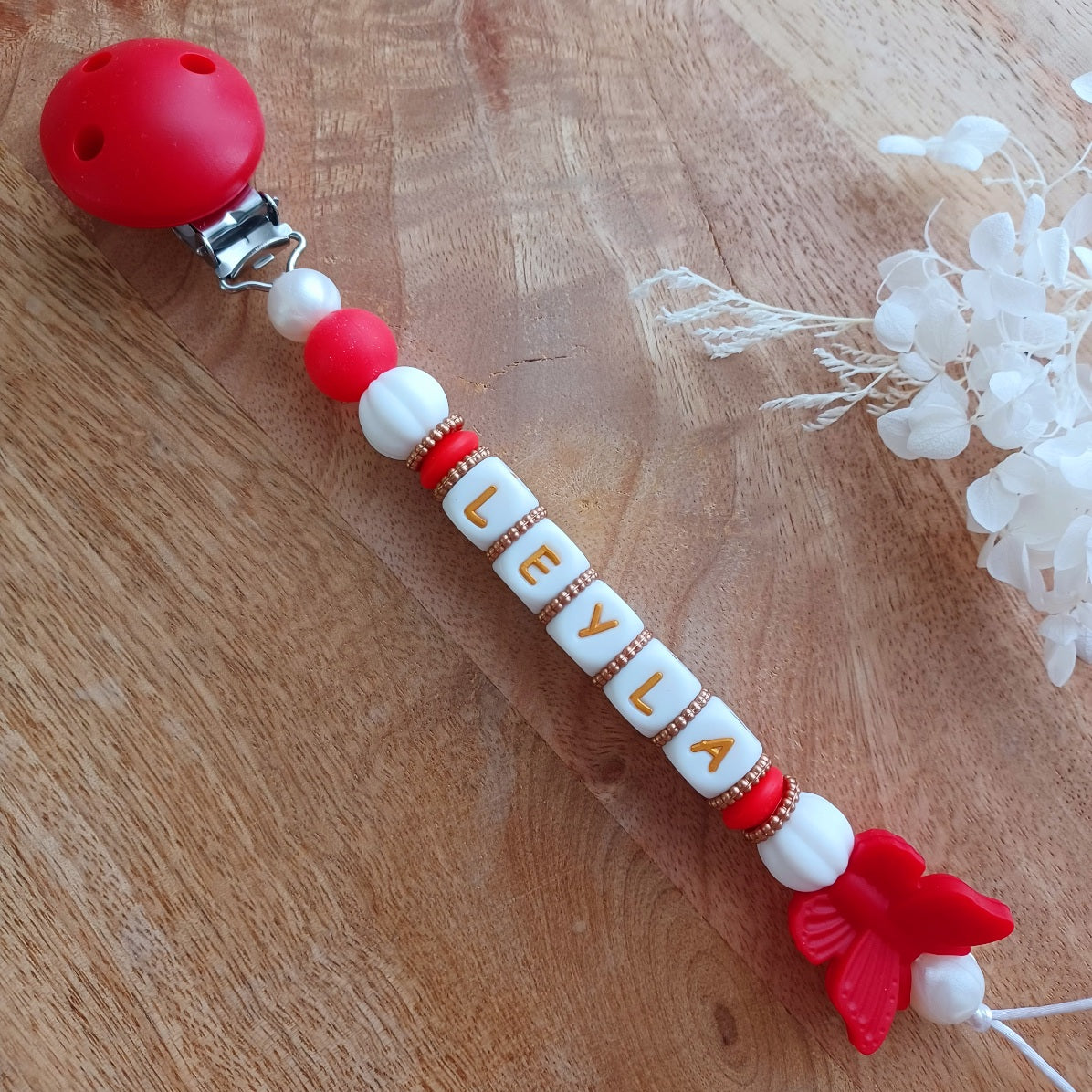 Attache tétine et anneau de dentition en silicone alimentaire sans BPA.  Clip rond en silicone et acier inoxydable, perles prénom personnalisable, papillon rouge en silicone. Anneau de dentition en taille parfaite pour les petites mains du bébé. Cadeau de naissance