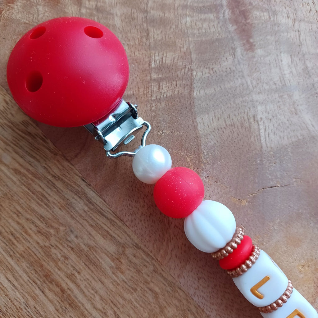 Attache tétine et anneau de dentition en silicone alimentaire sans BPA.  Clip rond en silicone et acier inoxydable, perles prénom personnalisable, papillon rouge en silicone. Anneau de dentition en taille parfaite pour les petites mains du bébé. Cadeau de naissance