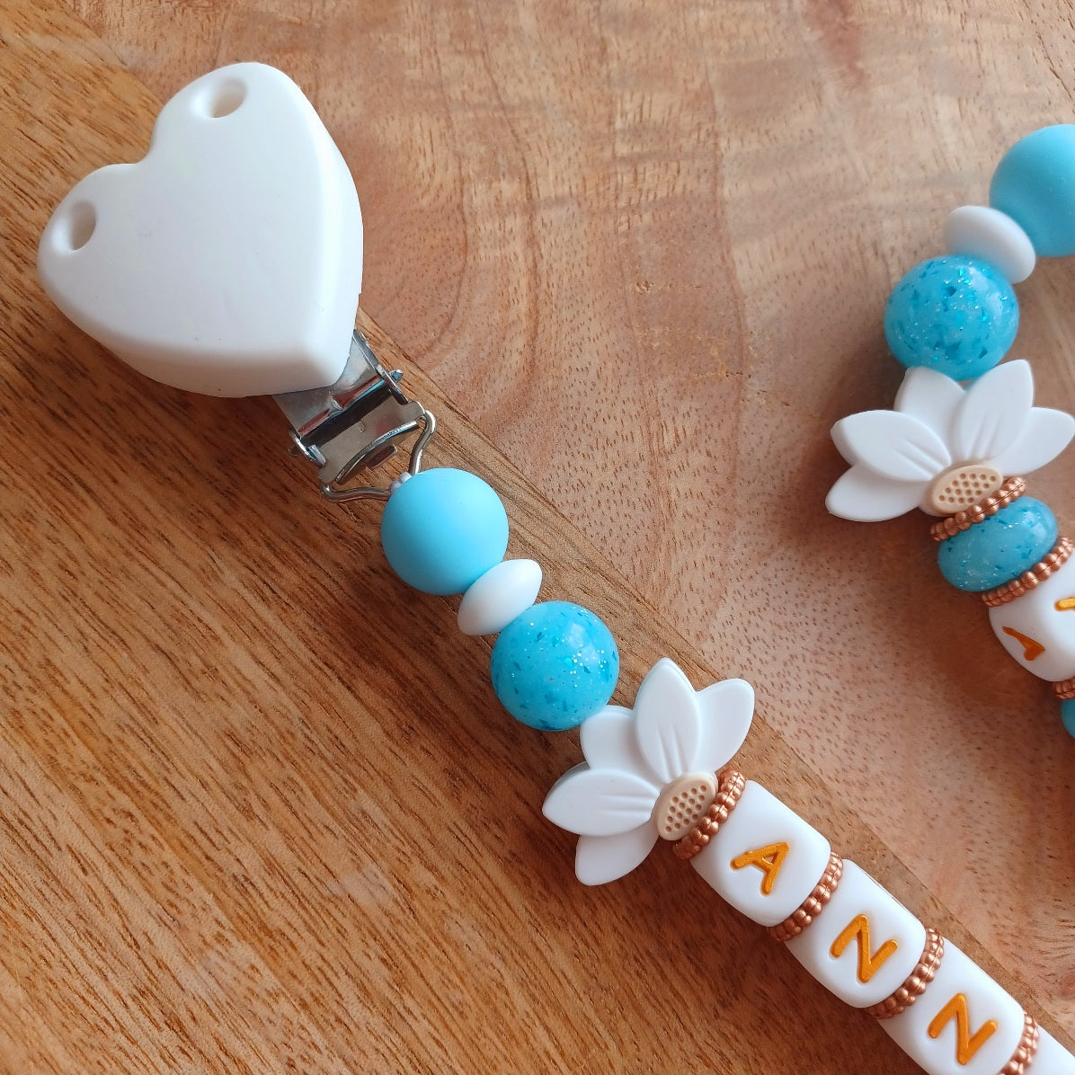 Attache tétine et anneau de dentition personnalisables avec le prénom de l'enfant. Réalisé en silicone alimentaire sans BPA, non toxique. Perles pailletées, perles en forme de fleur de lotus, clip en forme de coeur, pince en acier inoxydable. Super idée cadeau, cadeau de naissance. Couleur bleu et blanc, lettre en couleur blanc et doré.