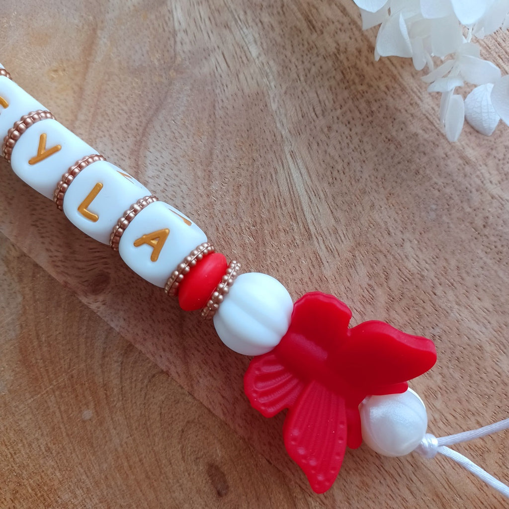 Attache tétine et anneau de dentition en silicone alimentaire sans BPA.  Clip rond en silicone et acier inoxydable, perles prénom personnalisable, papillon rouge en silicone. Anneau de dentition en taille parfaite pour les petites mains du bébé. Cadeau de naissance