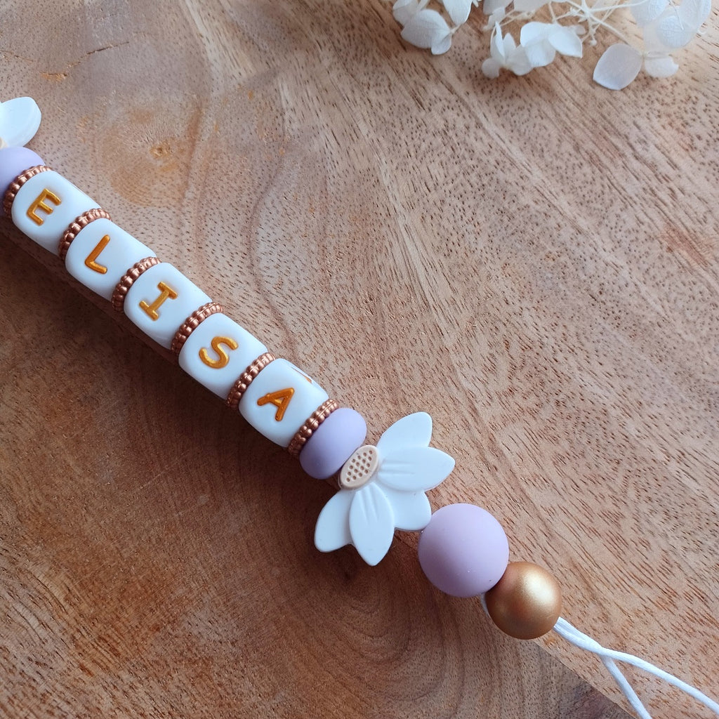 Attache tetine personnalisable, avec prénom du bébé ou enfant. Clip forme de coeur en silicone et acier inoxydable, perles du prénom en blanc et doré, perles rondes en lilas, perles de fleur de lotus. Cadeau de naissance