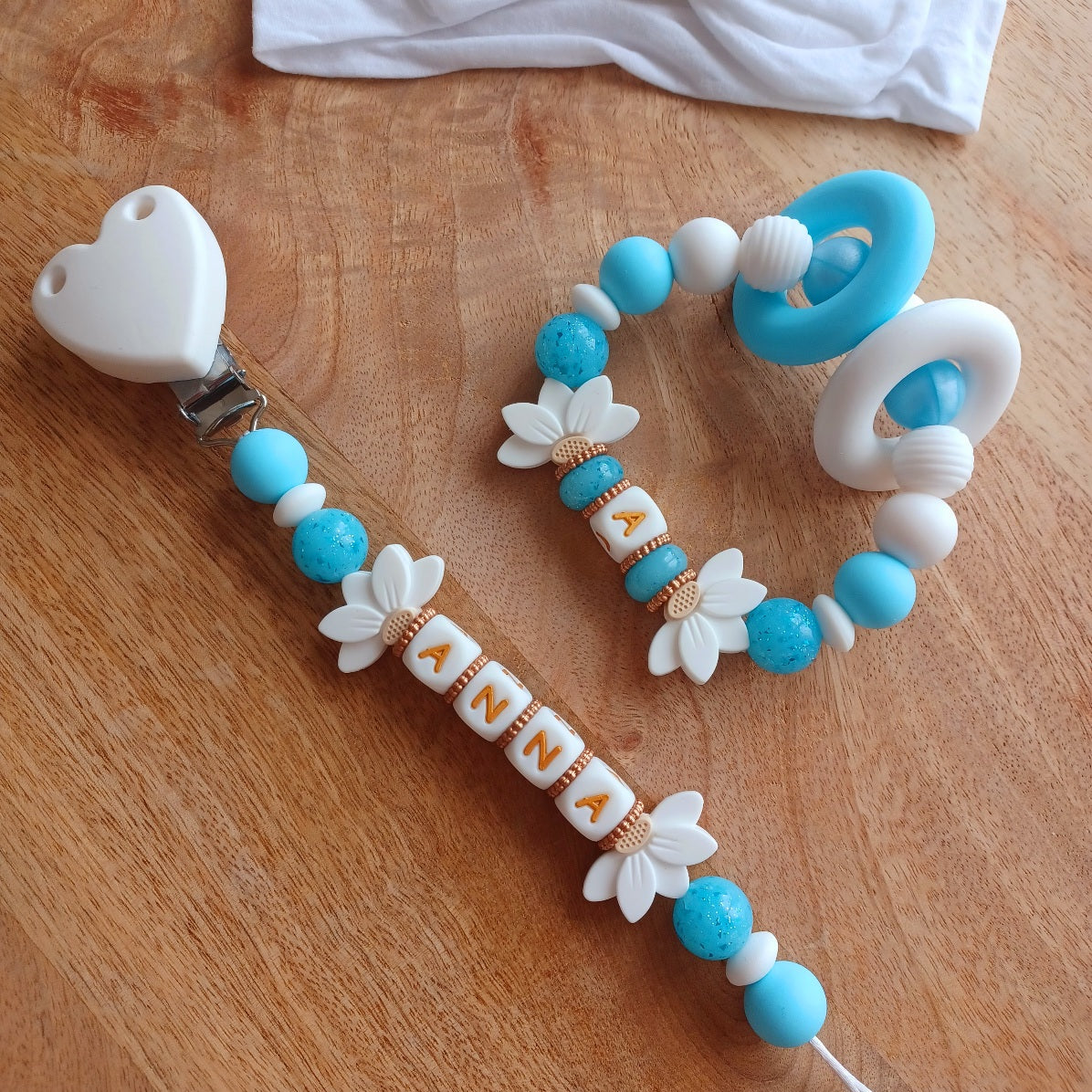 Attache tétine et anneau de dentition personnalisables avec le prénom de l'enfant. Réalisé en silicone alimentaire sans BPA, non toxique. Perles pailletées, perles en forme de fleur de lotus, clip en forme de coeur, pince en acier inoxydable. Super idée cadeau, cadeau de naissance. Couleur bleu et blanc, lettre en couleur blanc et doré.
