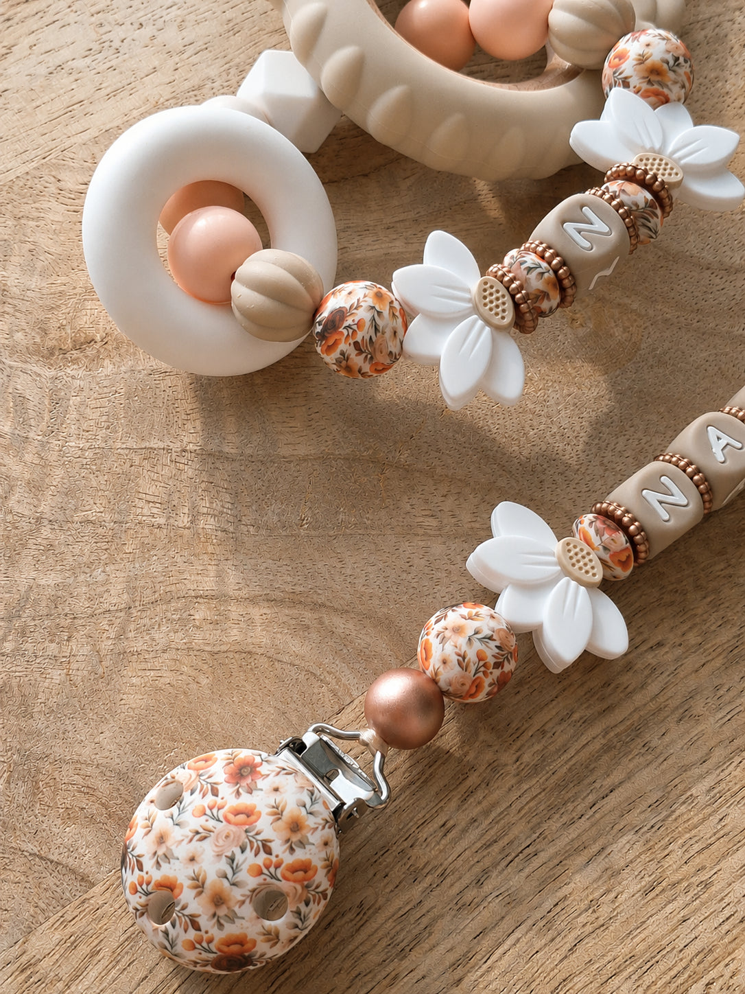 Attache tétine personnalisable avec prénom de l'enfant, en silicone alimentaire sans BPA. Clip robuste en silicone et acier inoxydable, perles imprimées, fleurs de lotus blanches. Anneau de dentition assorti.