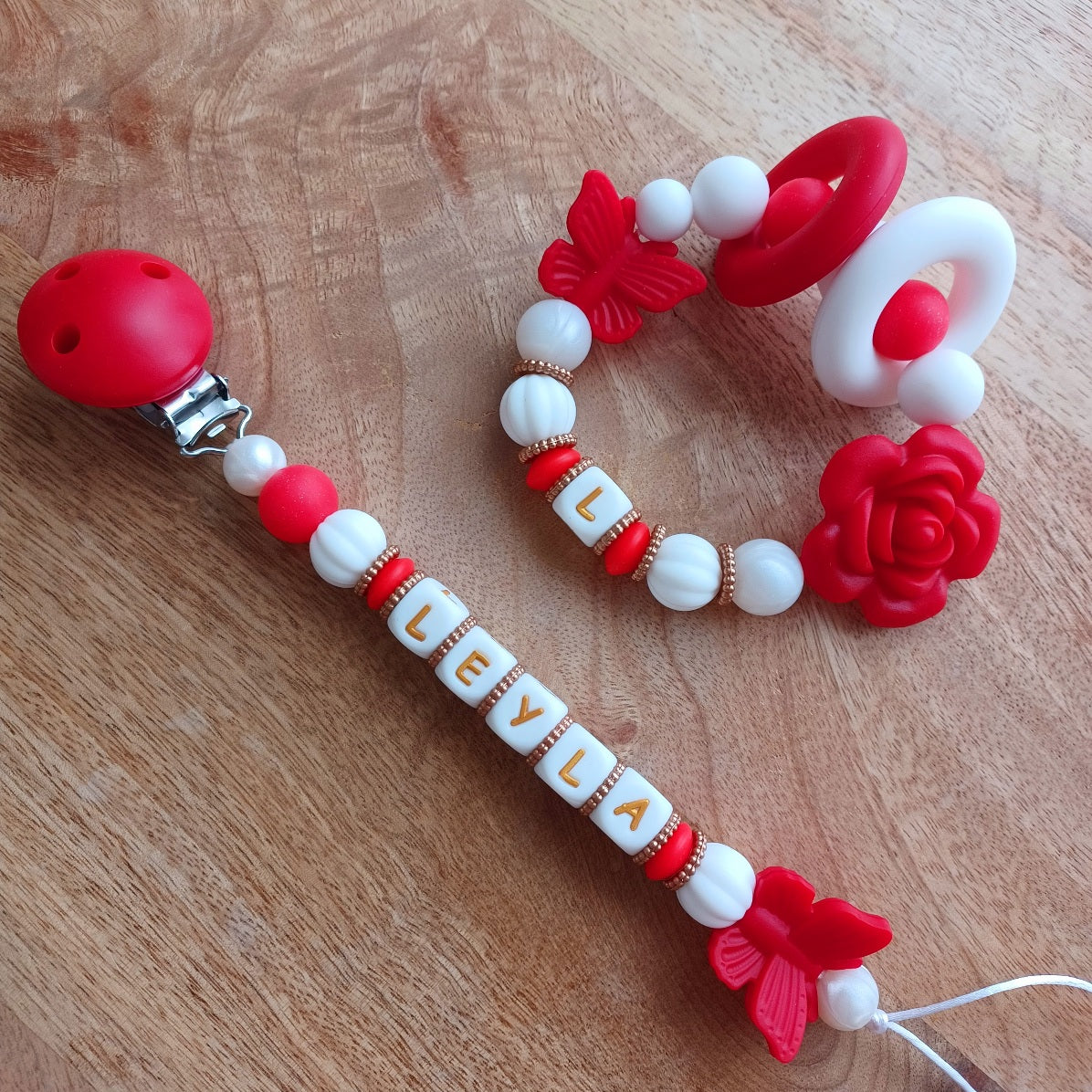 Attache tétine et anneau de dentition en silicone alimentaire sans BPA.  Clip rond en silicone et acier inoxydable, perles prénom personnalisable, papillon rouge en silicone. Anneau de dentition en taille parfaite pour les petites mains du bébé. Cadeau de naissance