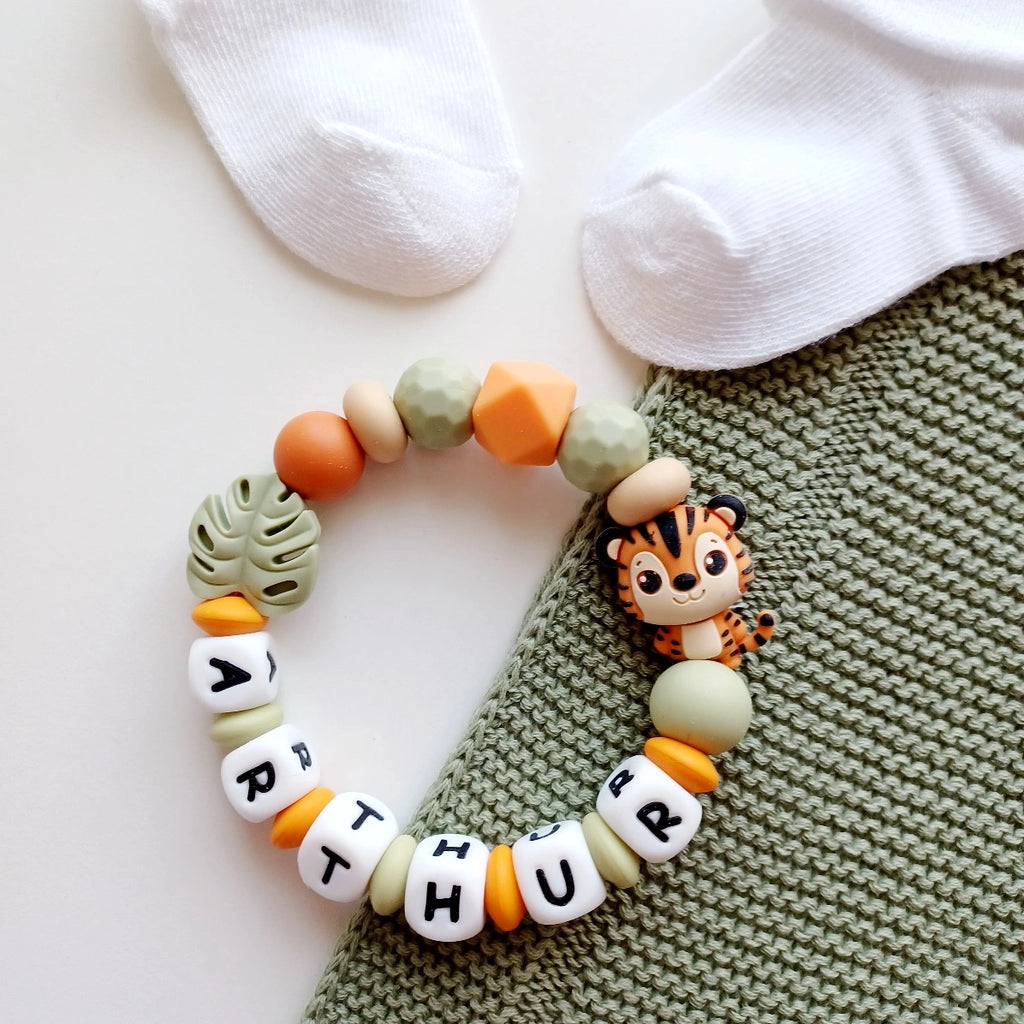 Anneau de dentition pour soulager les gencives des bébés. Cet anneau de dentition en forme de bracelet, est En silicone alimentaire sans BPA, entièrement personnalisables, perles vertes, beiges, oranges perles du prénom et perles fantaisie en forme de tigre t et feuille. Idéal cadeau naissance.