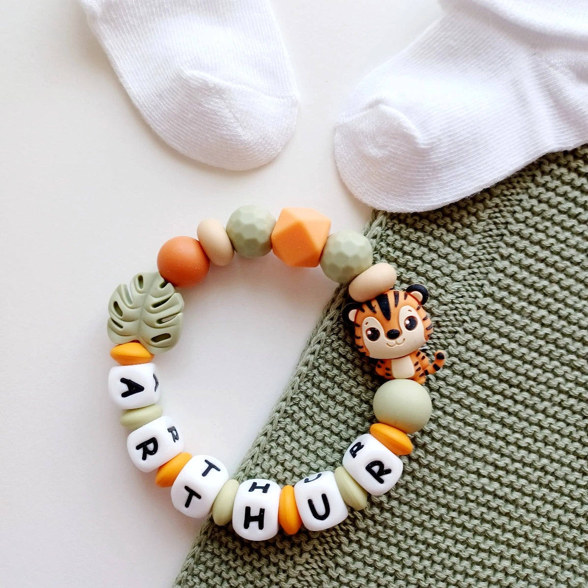 Anneau de dentition pour soulager les gencives des bébés. Cet anneau de dentition en forme de bracelet, est En silicone alimentaire sans BPA, entièrement personnalisables, perles vertes, beiges, oranges perles du prénom et perles fantaisie en forme de tigre t et feuille. Idéal cadeau naissance.